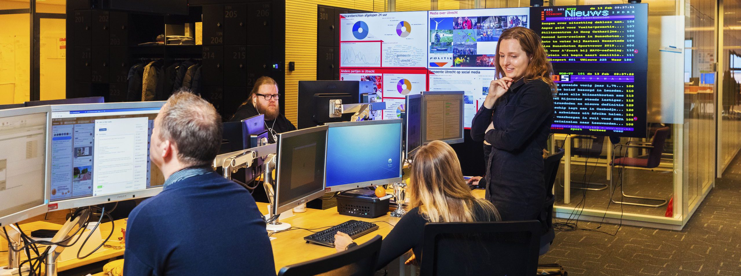 vier collega’s die zitten en staan om een blok van zes bureaus bij communicatie van gemeente Utrecht. Op de achtergrond zijn verschillende beeldschermen waar al het nieuws wordt getoond en bijhegouden.