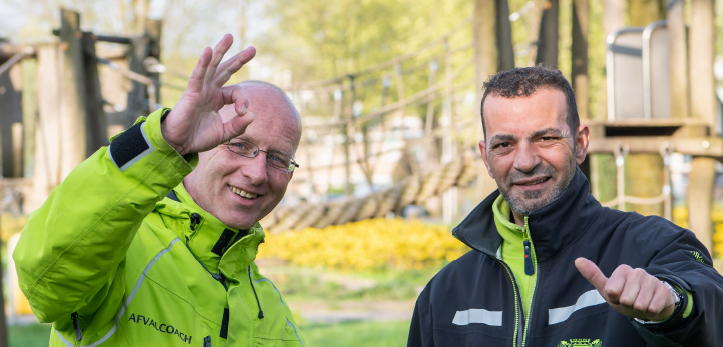 Twee collega’s staan bij een speeltuin en steken hun duim op. Op de achtergrond staat een houten speeltoestel met twee houten plateaus en daartussen een brug van touw. In de foto staat de tekst: “Door te snappen waarom mensen afval dumpen, kun je beter met ze in gesprek gaan”.