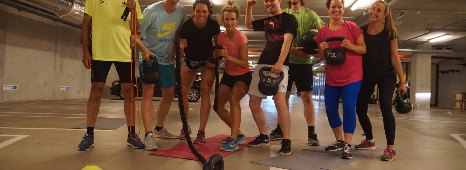 acht collega’s tijdens een bootcamp les die fit blijven in de parkeergarage van het stadskantoor met sport materialen zoals matjes, kettle bells staan.