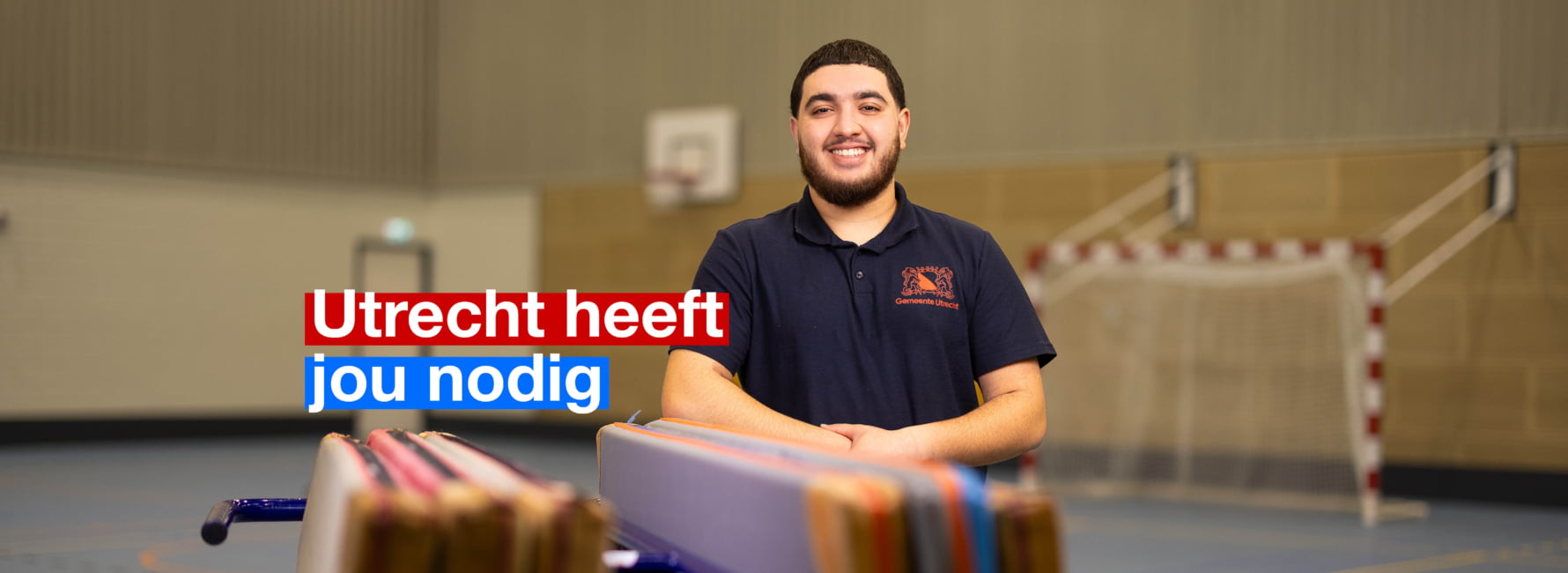 Beheerder Binnensport staat geleund tegen een stapel matten in een grote gymzaal. In beeld staat de tekst 'Utrecht heeft jou nodig'.