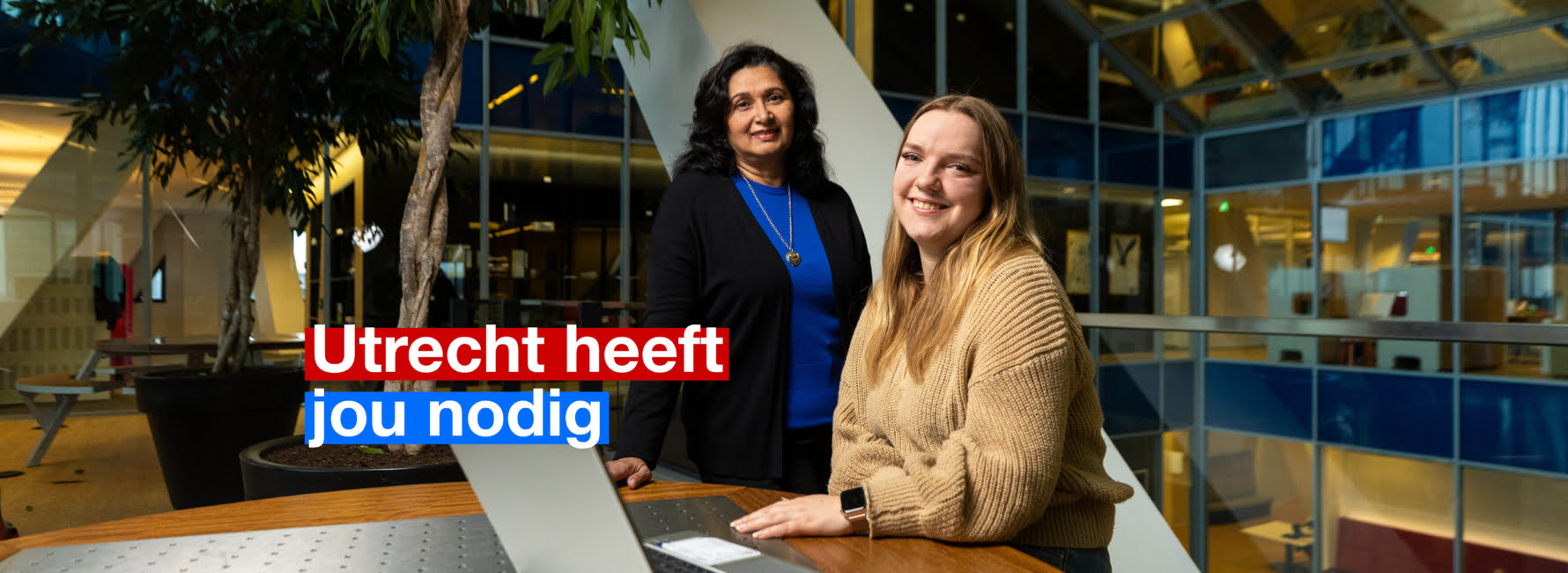 Twee personen. Eén persoon zit achter de laptop terwijl de andere daarachter staat. In beeld staat de tekst 'Utrecht heeft jou nodig'.