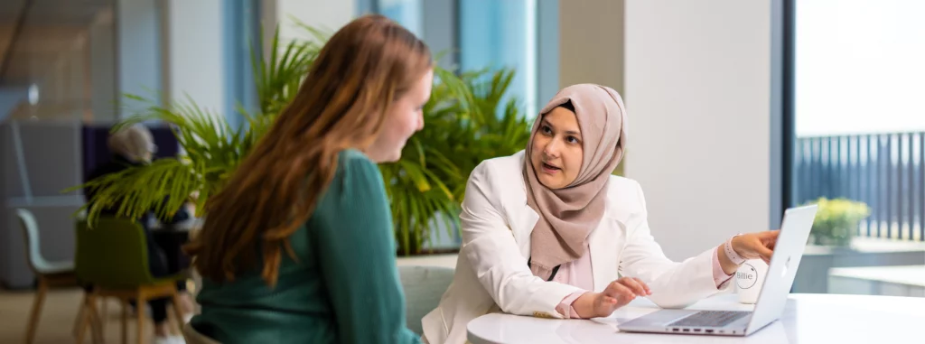Twee vrouwelijke medewerkers zitten aan een tafel en kijken naar een laptop